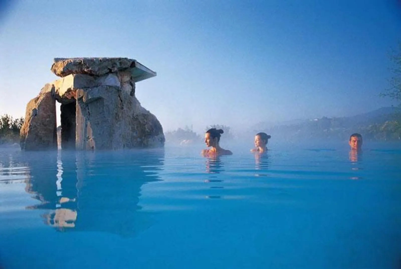 Piscina termale all'aperto con un dolmen antico, tre persone in acqua tra il vapore.